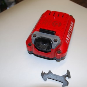 May include: A red Craftsman power tool battery with a black and gray connector. A gray metal tool is lying on a white surface.