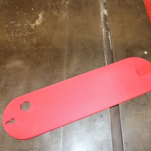 May include: A bright red table saw blade guard, oval in shape, with a rectangular tab on one end and a hole and keyhole shape on the other. The guard is lying on a wooden surface.