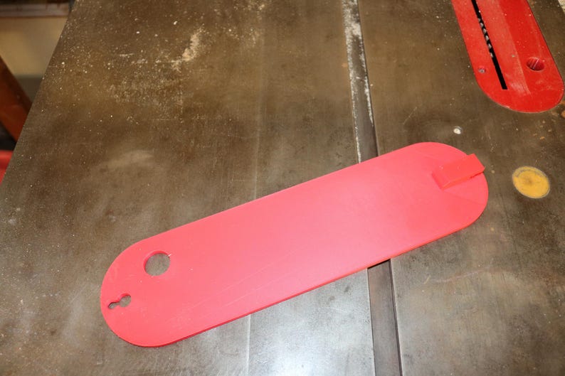 May include: A bright red table saw blade guard, oval in shape, with a rectangular tab on one end and a hole and keyhole shape on the other. The guard is lying on a wooden surface.