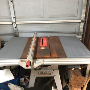 May include: A Ridgid table saw with a gray and brown tabletop and a red safety guard. The saw has a white base and is set up in a garage. The saw blade is visible on the left side.