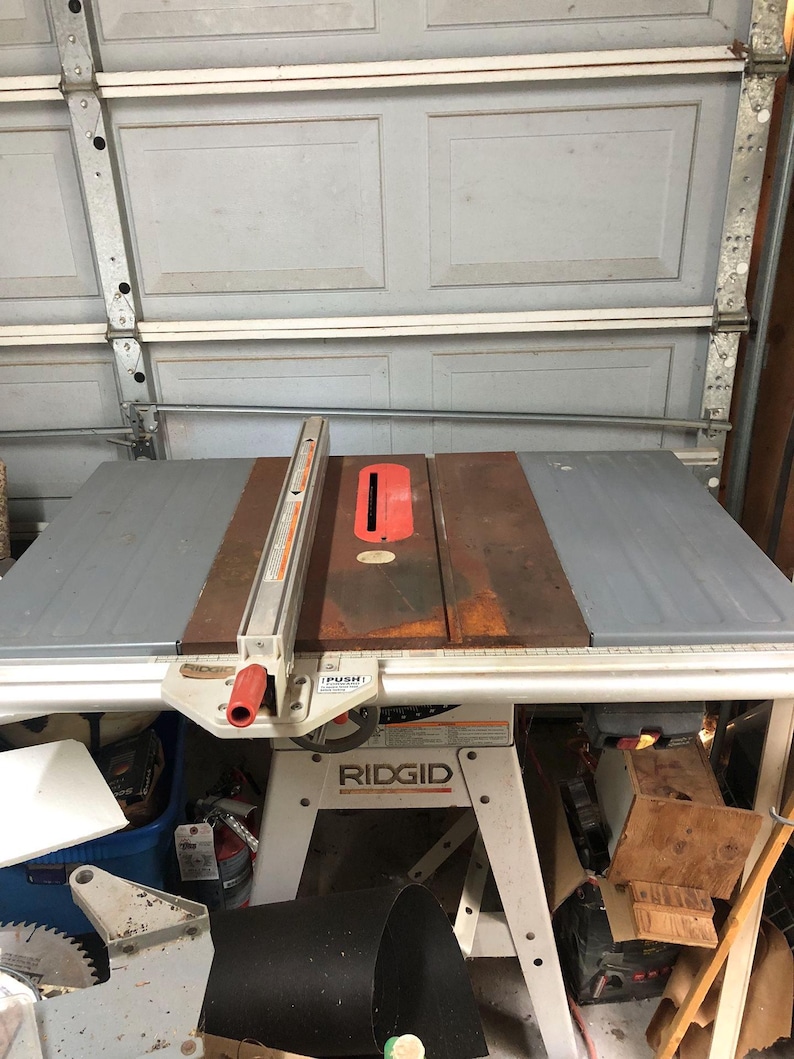 May include: A Ridgid table saw with a gray and brown tabletop and a red safety guard. The saw has a white base and is set up in a garage. The saw blade is visible on the left side.