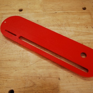 May include: A red plastic template with a long slot and two holes. The template is designed for woodworking projects.