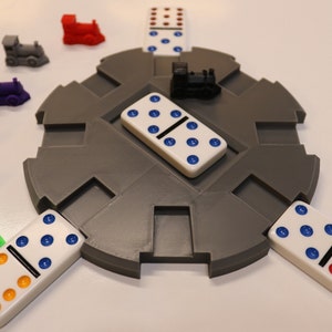 May include: A gray circular game board with five dominoes placed around the center. The dominoes have white backgrounds with blue and red dots. There are five small toy trains in different colors around the board.