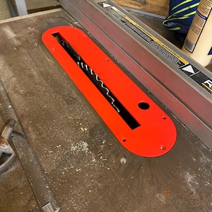 May include: A bright red table saw throat plate with a black blade opening. The plate is rectangular with rounded ends and has several screw holes. It sits on a wooden table saw surface.