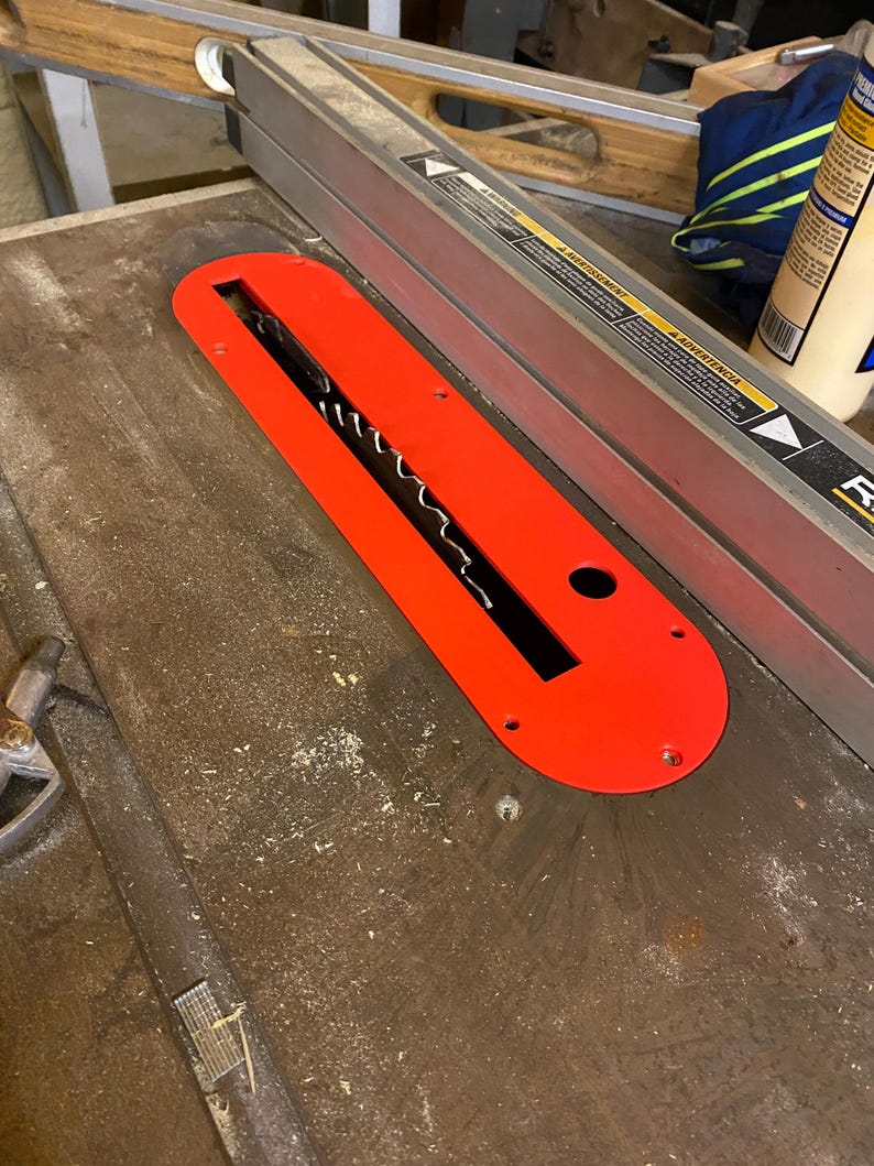 May include: A bright red table saw throat plate with a black blade opening. The plate is rectangular with rounded ends and has several screw holes. It sits on a wooden table saw surface.
