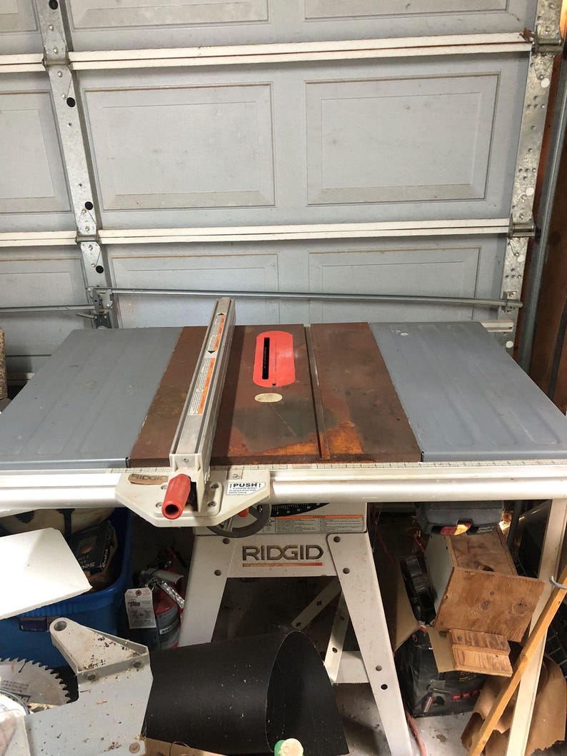 May include: A white and red Ridgid table saw with a gray table top and a metal fence. The saw is on a set of white metal legs. The saw is in a garage setting with other tools and equipment visible in the background.