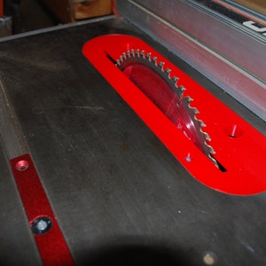 May include: A close-up of a table saw blade with a red safety guard. The blade is partially visible and has a serrated edge. The guard is made of metal and has a red finish.
