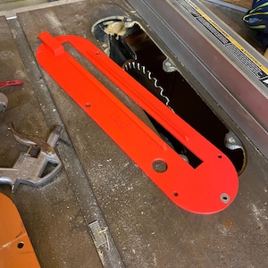 May include: A bright red table saw blade guard with a rectangular shape and several holes. The guard is positioned over a circular saw blade, with the blade's teeth visible. The saw is on a metal surface.
