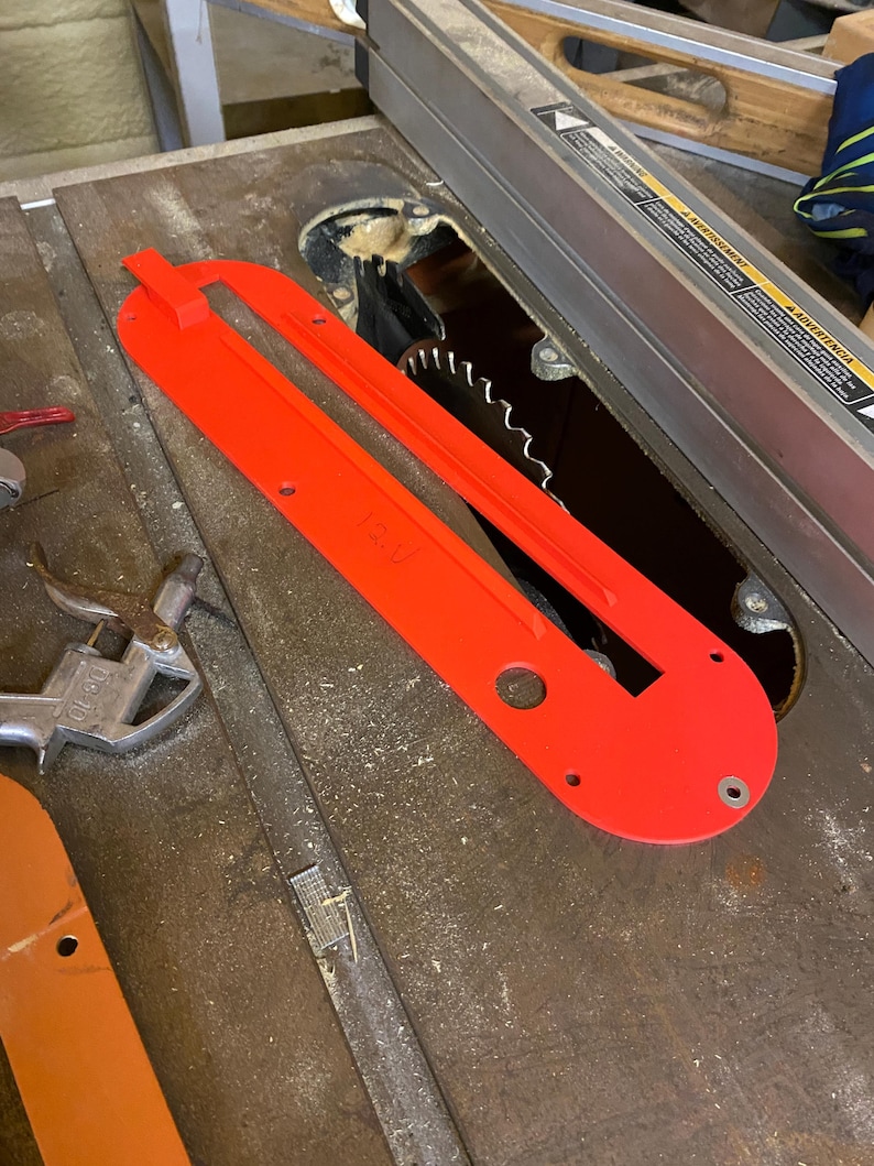 May include: A bright red table saw blade guard with a rectangular shape and several holes. The guard is positioned over a circular saw blade, with the blade's teeth visible. The saw is on a metal surface.