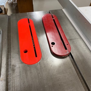 May include: Two red plastic table saw inserts with a slot in the middle. The inserts are on a metal table saw surface.