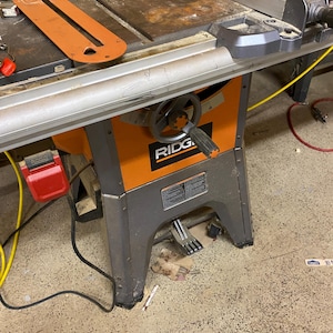 May include: An orange and gray table saw with a red blade guard. The saw has a silver table and a black handle. The brand name "RIDGID" is visible on the front of the saw.