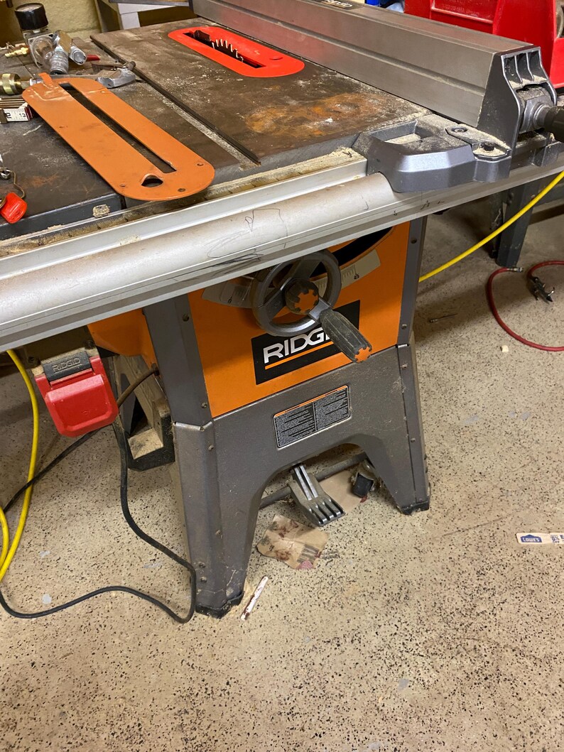 May include: An orange and gray table saw with a red blade guard. The saw has a silver table and a black handle. The brand name "RIDGID" is visible on the front of the saw.