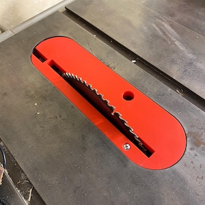 May include: A close-up of a table saw with a red blade guard. The circular saw blade is visible, with sharp teeth. The saw table is gray and has a metal surface. A small piece of metal is in the lower left corner.