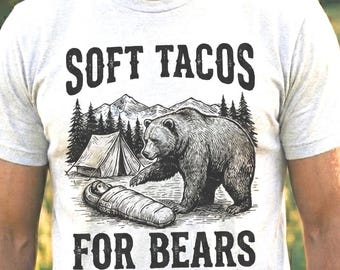 Puede incluir: Una camiseta gris claro con el texto "SOFT TACOS FOR BEARS" en negrita negra. El gráfico representa un oso cerca de una tienda de campaña y un saco de dormir. La camiseta la lleva una persona con barba, en un entorno natural al aire libre.