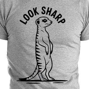 Peut inclure: T-shirt gris avec une illustration noire d'un suricate debout. Le texte "LOOK SHARP" est inscrit au-dessus. Le t-shirt est à col rond et à manches courtes.