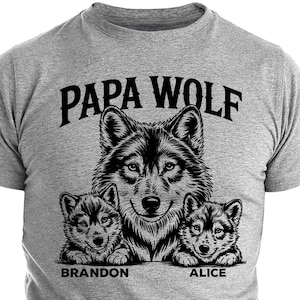 May include: Gray t-shirt featuring a black graphic of a wolf family. The design includes a large wolf and two smaller wolf pups, with the text "PAPA WOLF" above and the names "BRANDON" and "ALICE" below the pups.