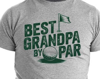 Puede incluir: Una camiseta gris con el texto verde "BEST GRANDPA BY PAR", una pelota de golf y un gráfico de bandera. La camiseta está hecha de un material suave y tiene mangas cortas.