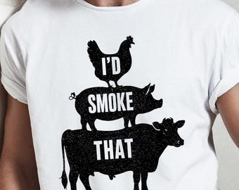 May include: White t-shirt with a black graphic of a chicken, pig, and cow stacked on top of each other. The text "I'D SMOKE THAT" is printed on the shirt in a bold, sans-serif font. The shirt is made of a soft material.