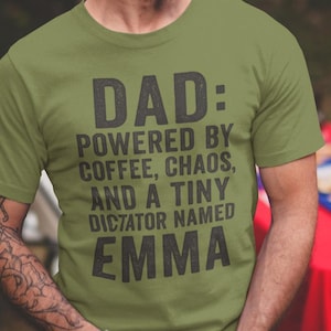 以下が含まれることがあります： オリーブグリーンのTシャツ。黒い文字で「DAD: POWERED BY COFFEE, CHAOS, AND A TINY DICTATOR NAMED EMMA」と書かれています。ひげを生やした男性がTシャツを着て笑顔です。背景にはお祝いの飾りがあります。