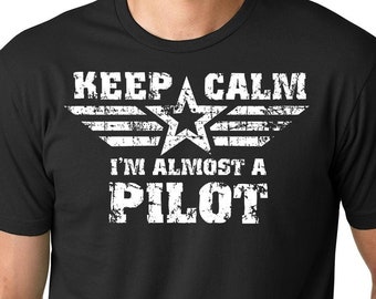 May include: Black t-shirt with a white distressed graphic that says "Keep Calm I'm Almost A Pilot" with a star and wings.