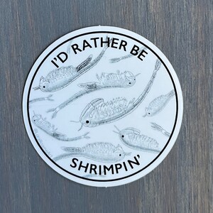 May include: A round white sticker with the text "I'D RATHER BE SHRIMPING'" in black. The sticker features a detailed illustration of several shrimp-like creatures in a circular pattern. The background is a gray wooden surface.