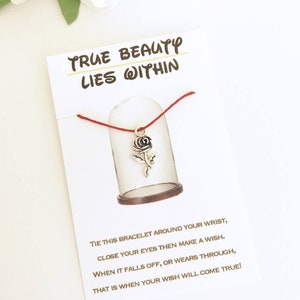 May include: A card with the text "TRUE BEAUTY LIES WITHIN" and a red string bracelet with a silver rose charm. The card includes instructions on how to wear the bracelet and make a wish.