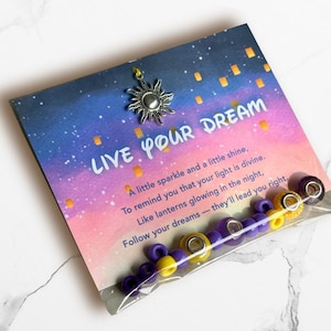 May include: A jewelry-making kit with a card that reads "LIVE YOUR DREAM." The card has a starry sky design with floating lanterns. The kit includes purple and yellow beads, and a silver sun charm.