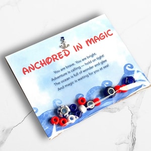 May include: A card with the words "ANCHORED IN MAGIC" and a poem about adventure. The card is decorated with a small silver anchor charm and a string of red, white, and blue beads.