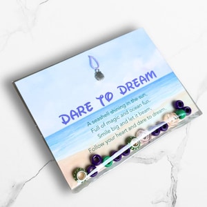 May include: A package with the words "DARE TO DREAM" on a blue and white background. The package contains beads in various colors, including green, purple, and clear. The text on the package reads "A seashell shining in the sun, Full of magic and ocean fun."