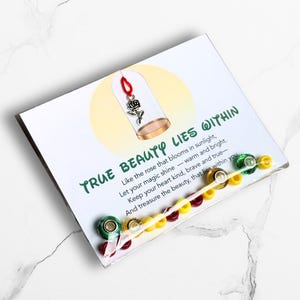 May include: A card with the text "TRUE BEAUTY LIES WITHIN" and a rose charm inside a glass dome. The card has a clear plastic strip with colorful beads attached. The background is white with a marble pattern.