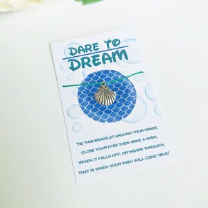 Könnte beinhalten: Eine weiße Karte mit dem Text "DARE TO DREAM" in Türkis. Ein türkisfarbenes Armband mit einem silbernen Muschel-Anhänger ist an der Karte befestigt. Die Karte hat ein blaues Schuppenmuster und den Text "TIE THIS BRACELET..." und "THAT IS WHEN YOUR WISH WILL COME TRUE!"
