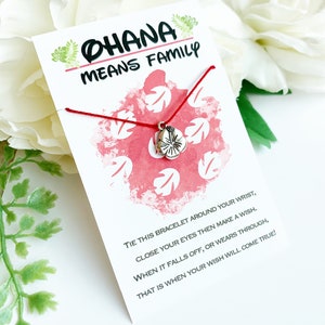 K&ouml;nnte beinhalten: Eine wei&szlig;e Karte mit dem Text "OHANA MEANS FAMILY" und einem roten Schnurarmband mit einem silbernen Blumenanh&auml;nger. Die Karte hat ein rot-wei&szlig;es Blumenmuster und Anweisungen.
