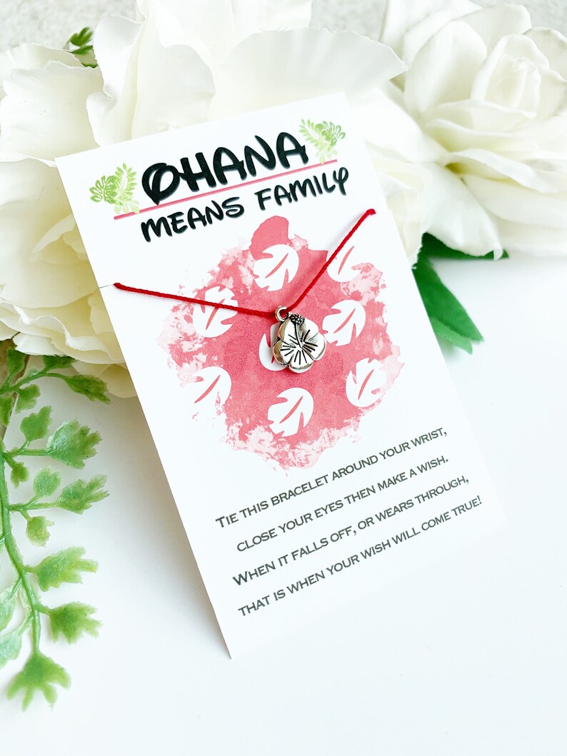 K&ouml;nnte beinhalten: Eine wei&szlig;e Karte mit dem Text "OHANA MEANS FAMILY" und einem roten Schnurarmband mit einem silbernen Blumenanh&auml;nger. Die Karte hat ein rot-wei&szlig;es Blumenmuster und Anweisungen.