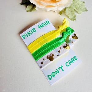 Op de afbeelding: Drie kleurrijke haar elastiekjes op een witte kaart met de tekst "PIXIE HAIR" en "DON'T CARE". De haar elastiekjes zijn geel, groen en wit. Het witte haar elastiekje heeft gouden Minnie Mouse hoofden en hartjes.