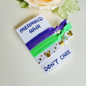 Puede incluir: Un conjunto de cuatro cintas para el pelo en morado, verde y blanco, con detalles dorados. La cinta blanca presenta cabezas y corazones dorados de Minnie Mouse. La tarjeta dice "MERMAID HAIR" y "DON'T CARE" en azul.