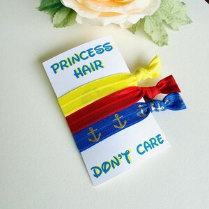 Op de afbeelding: Een set van drie haarbanden in geel, rood en blauw, met gouden ankeraccenten op de blauwe band. De kaart leest "PRINCESS HAIR" en "DON'T CARE" in blauwe en gele tekst. De haarbanden liggen op een witte kaart.