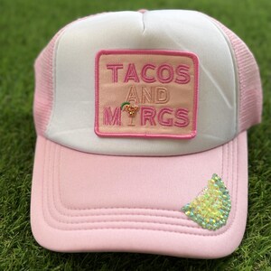 May include: A light pink and white trucker hat featuring a rectangular patch with the text TACOS AND MARGS, where the letter A in MARGS is replaced by a small cocktail glass icon. A lime wedge made of iridescent green sequins sits on the brim.
