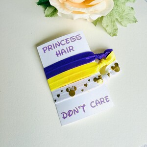 Op de afbeelding: Een set van vier haarbanden in paars, geel en wit. De witte haarband heeft gouden Mickey Mouse-hoofden en hartjes. De kaart leest "PRINCESS HAIR DON'T CARE" in paarse tekst.