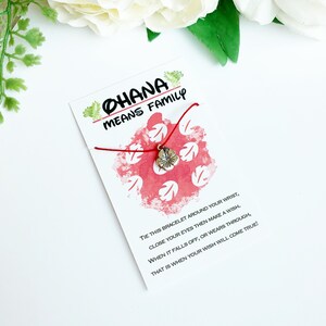 K&ouml;nnte beinhalten: Ein rotes Armband mit einem silbernen Blumenanh&auml;nger ist an einer Karte befestigt. Die Karte tr&auml;gt die Aufschrift "OHANA MEANS FAMILY" und ein rot-wei&szlig;es Design. Die Karte enth&auml;lt auch Anweisungen.