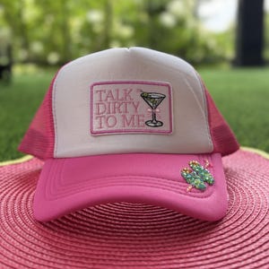 May include: A bright pink and white trucker hat featuring a rectangular patch with the text TALK DIRTY TO ME and an embroidered martini glass graphic. The brim is adorned with a cluster of iridescent green crystal embellishments.