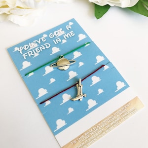 May include: Two friendship bracelets with charms on a blue background with white clouds. The charms are a silver boot and a silver planet. The text on the card reads "You've got a friend in me". Tie this bracelet around your wrist, close your eyes, make a wish. When it falls off or wears through, that is when your wish will come true.