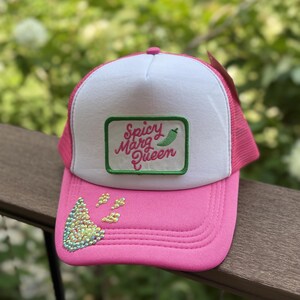 May include: A pink and white trucker hat featuring a rectangular patch with a green border and the text Spicy Marg Queen in pink script next to a green chilli pepper icon. The pink brim is decorated with iridescent rhinestones in a triangular shape.