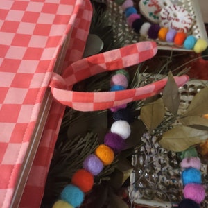 May include: A pink and white checkered book cover with a matching checkered strap. The book cover is lying on a table with a colorful garland of felt balls.