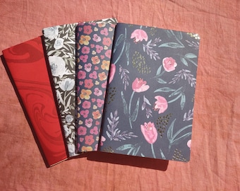 May include: Four different notebooks with floral patterns. The notebooks have a red, white and black floral pattern, a pink and orange floral pattern, a pink and blue floral pattern, and a red and white marbled pattern.
