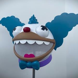 May include: A whimsical clown head sculpture with a white face, large blue hair, and a brown mouth. The clown has a red nose, black cross-shaped eyes, and a blue bow tie. The sculpture is mounted on a metal pole.