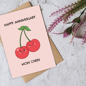 Puede incluir: Una tarjeta rosa con el texto "Happy Anniversary" y un dibujo de dos cerezas rojas con caras. El texto "Mon Ch&eacute;ri" est&aacute; debajo de las cerezas.