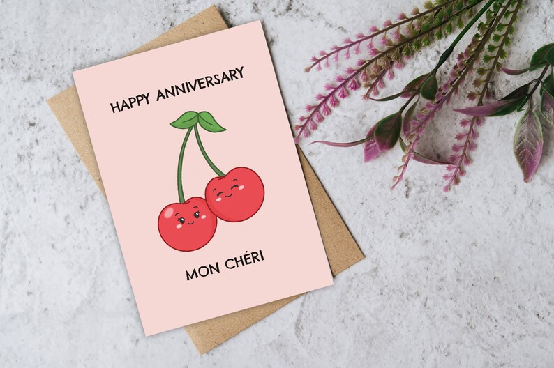 Puede incluir: Una tarjeta rosa con el texto "Happy Anniversary" y un dibujo de dos cerezas rojas con caras. El texto "Mon Ch&eacute;ri" est&aacute; debajo de las cerezas.