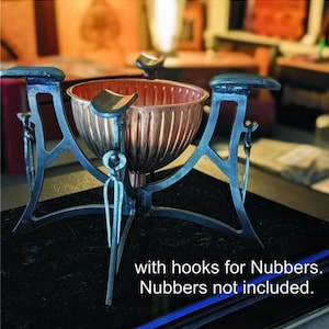 以下が含まれることがあります： 黒い金属製のスタンドに置かれた銅製のボウル。スタンドは3本の脚と各脚にフックが付いています。スタンドの下には「with hooks for Nubbers. Nubbers not included.」というテキストが印刷されています。