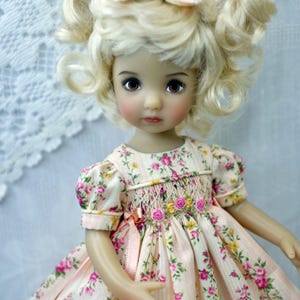 May include: A doll with blonde, curly hair and brown eyes, wearing a pink floral dress with a matching bow. The dress features a smocked bodice and pink ribbon accents, creating a vintage aesthetic.