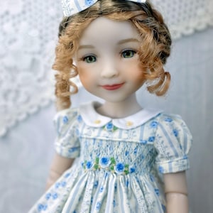 May include: A doll with green eyes and curly auburn hair, wearing a white and blue floral print dress with a matching bow. The dress has a white collar and a bodice with blue rose accents. The doll's face is delicately painted with rosy cheeks and red lips.